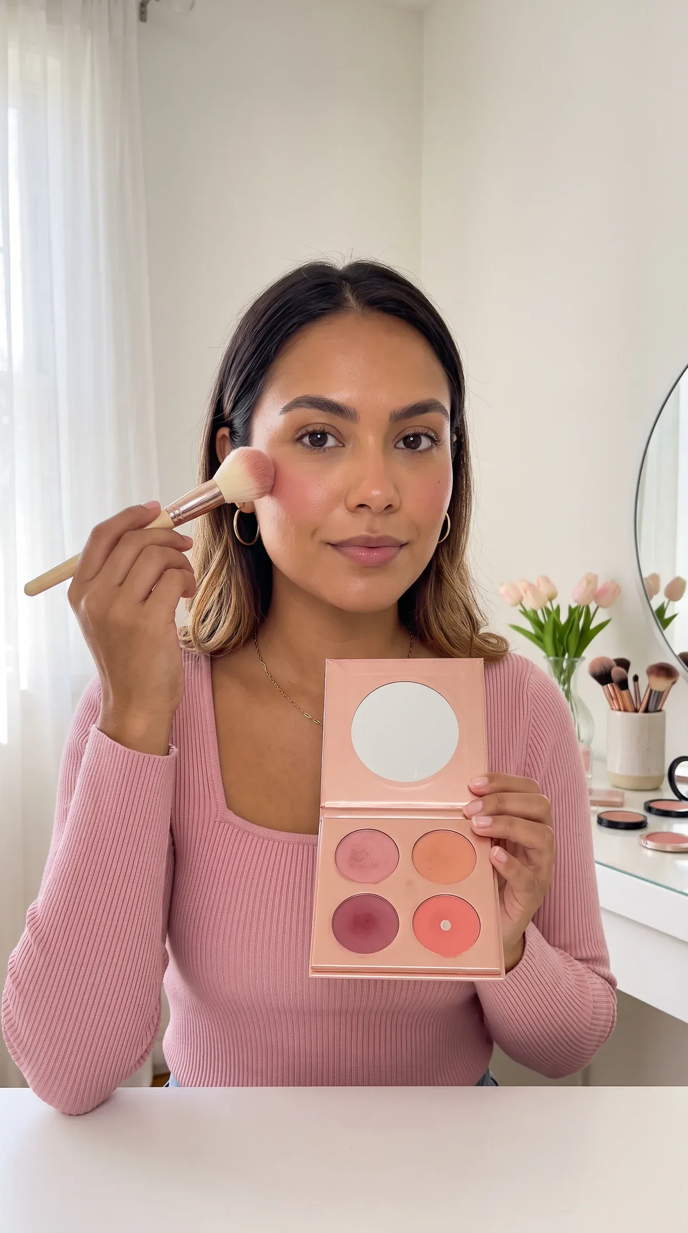 AI-generated persona in a pink ribbed top at a white vanity, holding an open four-shade blush palette toward the camera with a blush brush on her cheekbone
